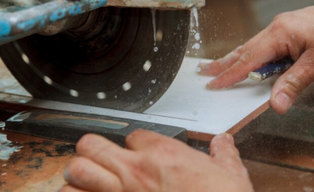 Close-up of a person's hand holding a ceramic piece as it moves through a circular saw. The saw drips with water.